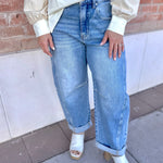Person wearing a white blouse, blue jeans, and white sandals standing against a brick wall.