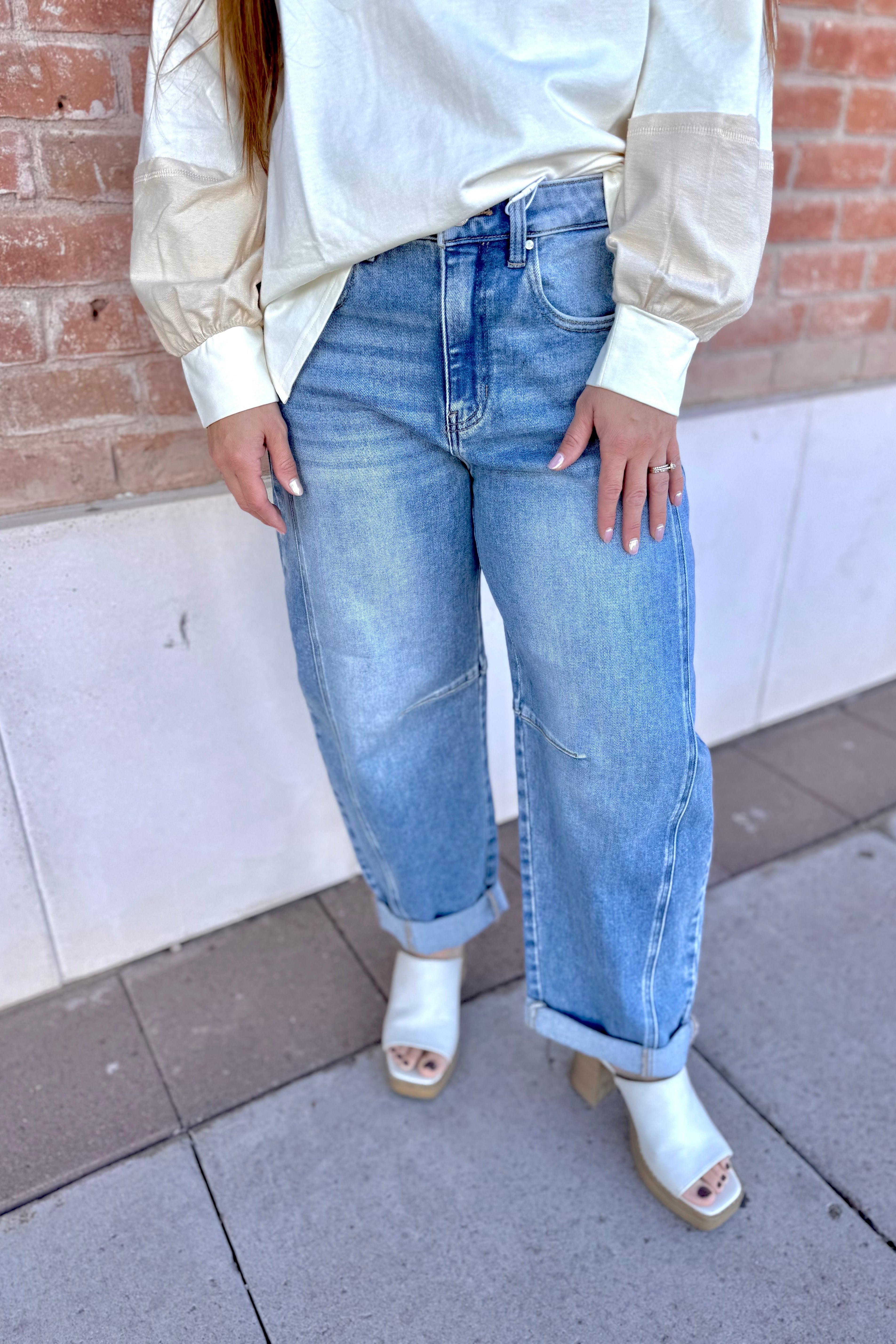 Person wearing a white blouse, blue jeans, and white sandals standing against a brick wall.