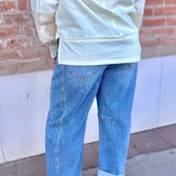 Person wearing a light-colored top and blue jeans standing against a brick wall.