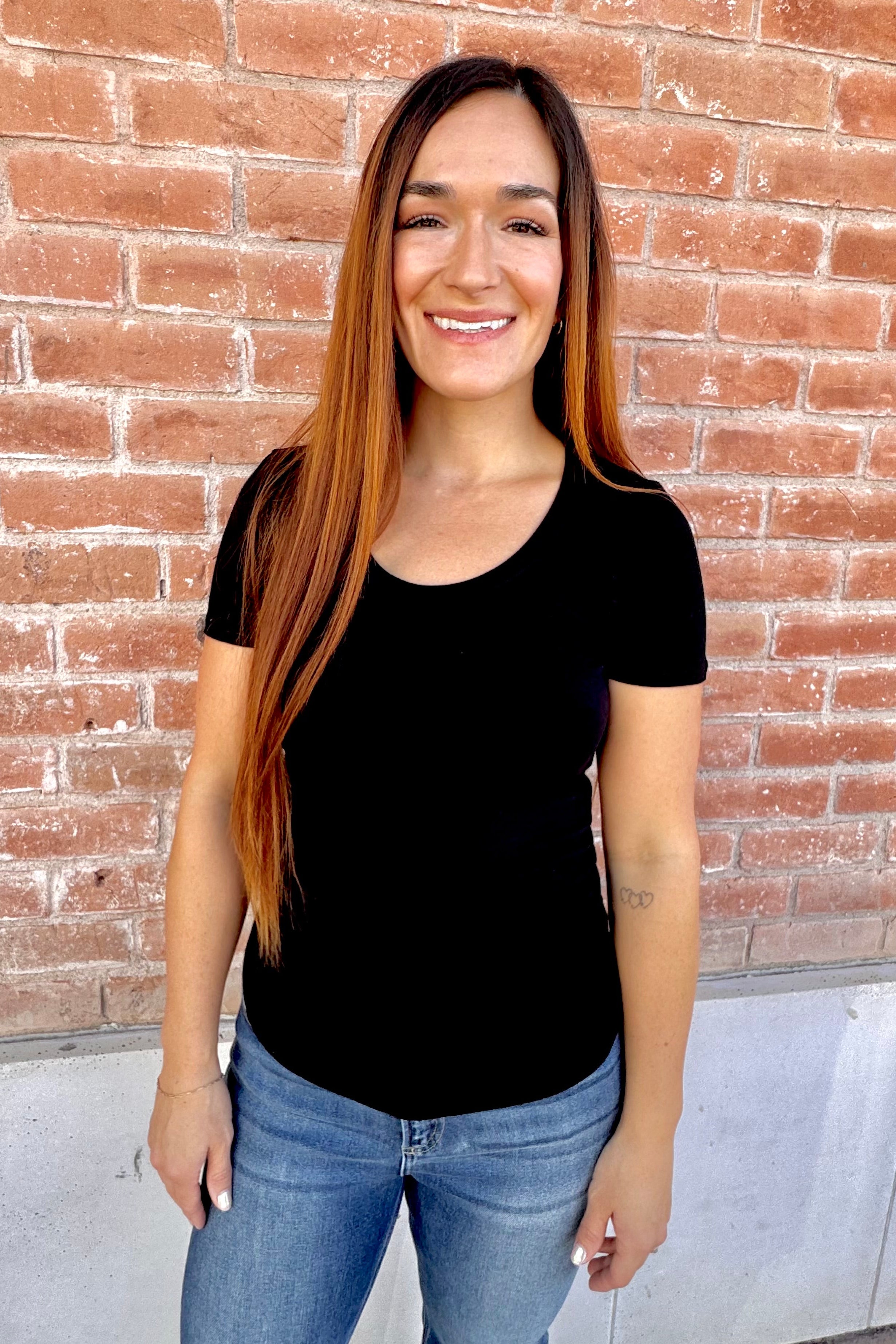 Woman wearing a black t-shirt and blue jeans standing in front of a brick wall.