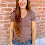 Woman wearing a brown t-shirt and blue jeans standing against a brick wall.