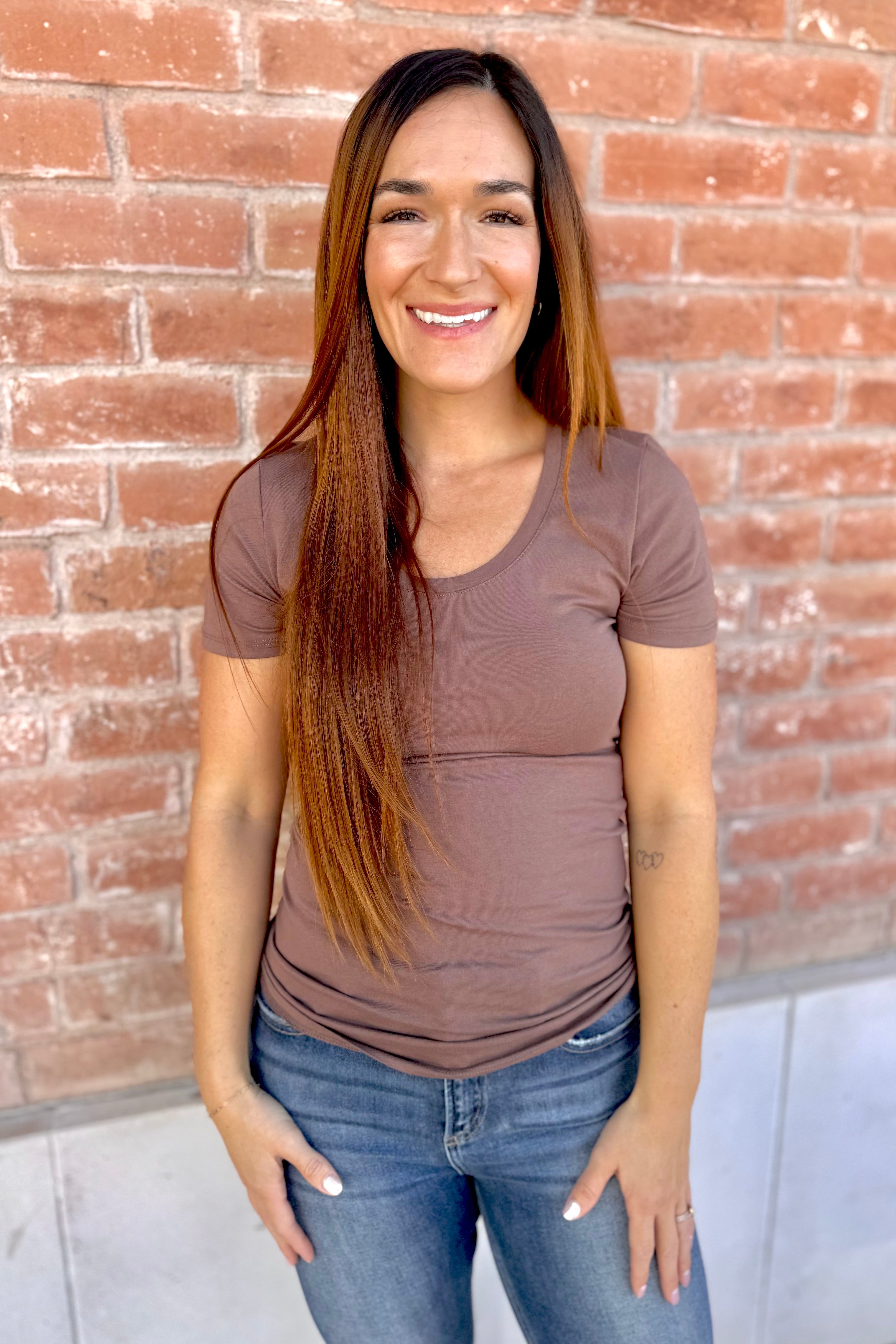 Woman wearing a brown t-shirt and blue jeans standing against a brick wall.