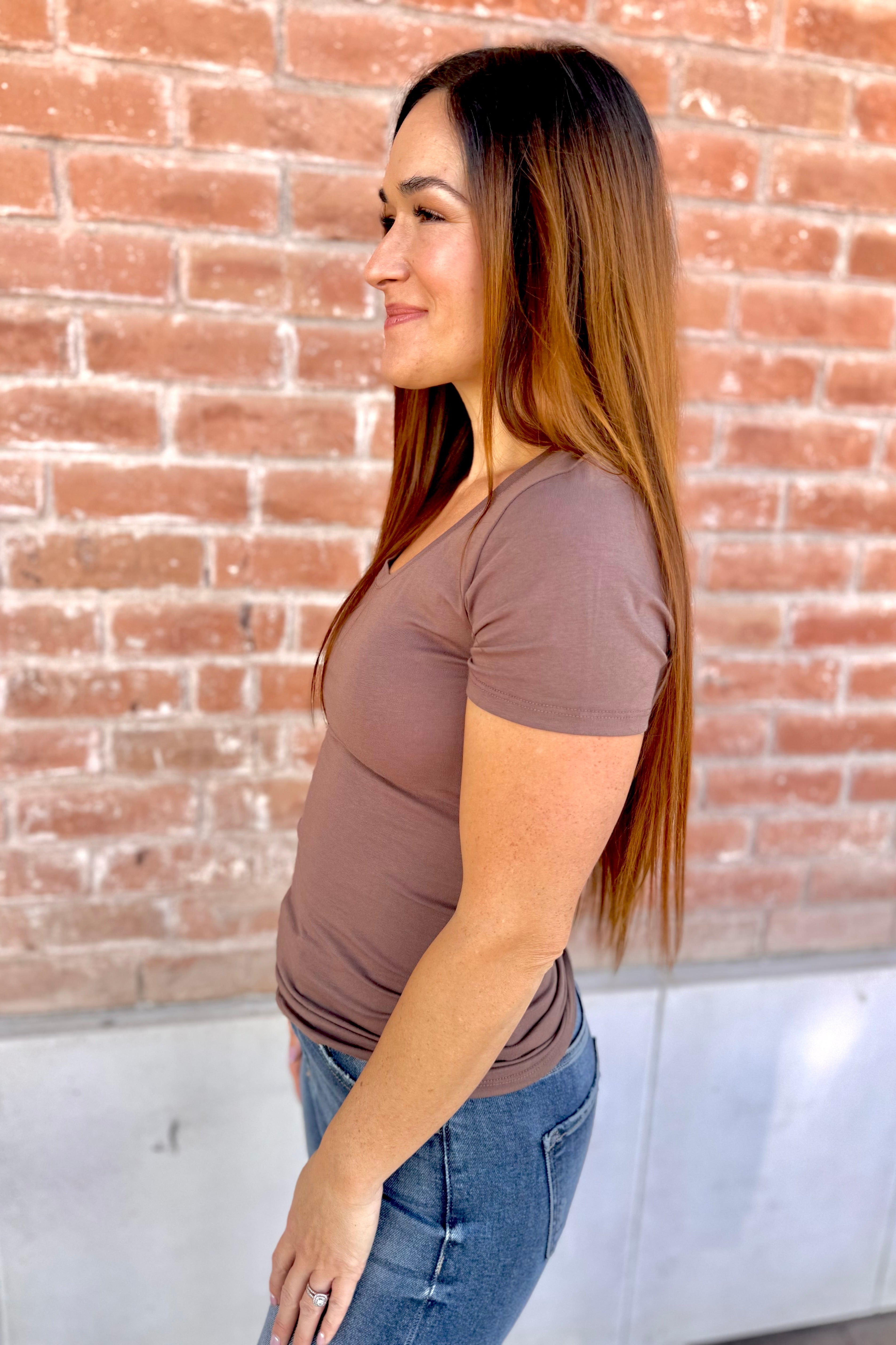Woman wearing a brown t-shirt and blue jeans standing against a brick wall.
