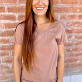 Woman wearing a brown t-shirt and blue jeans standing against a brick wall.