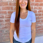 Woman wearing a light purple t-shirt and jeans standing in front of a brick wall.