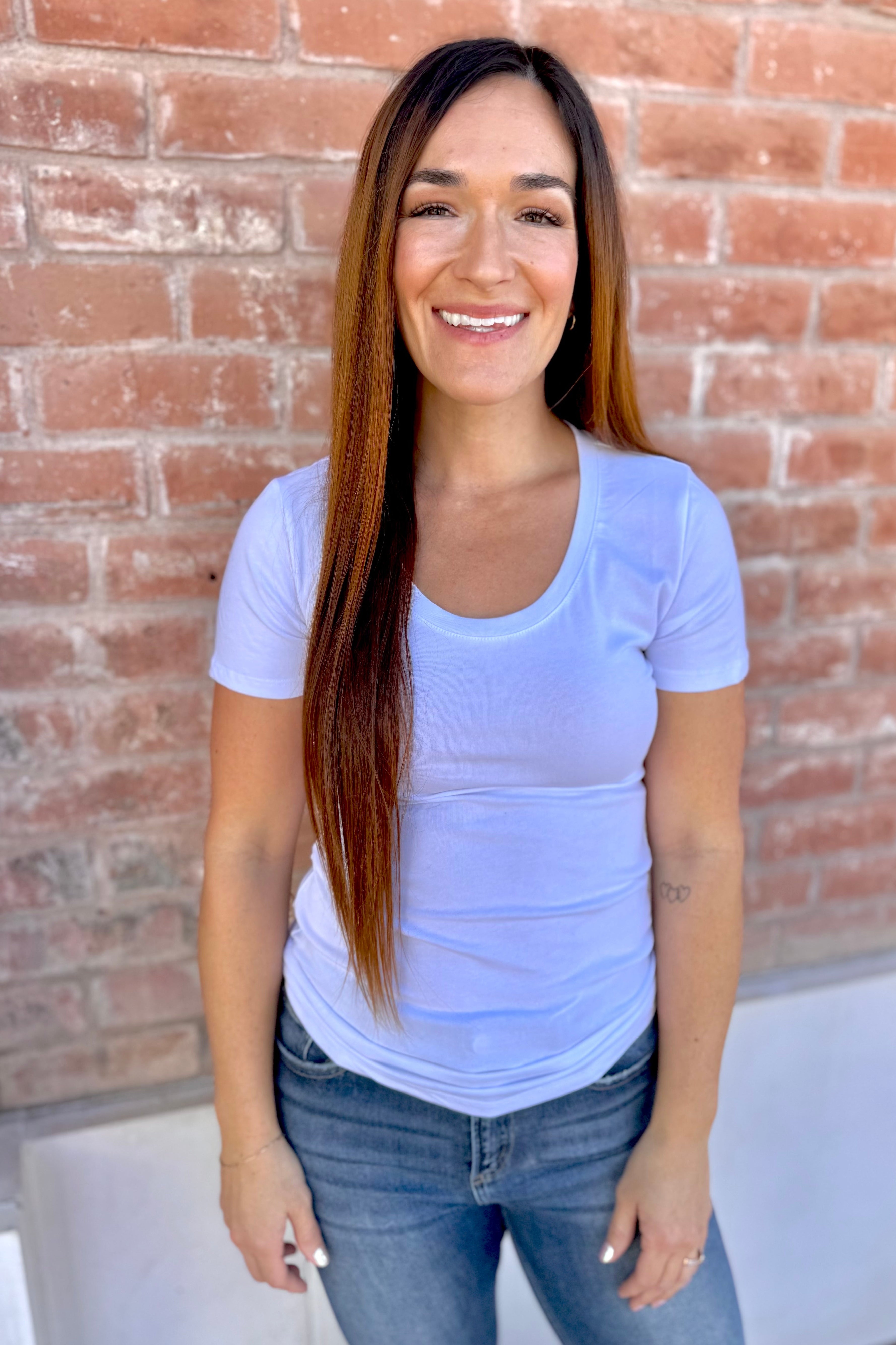 Woman wearing a light purple t-shirt and jeans standing in front of a brick wall.