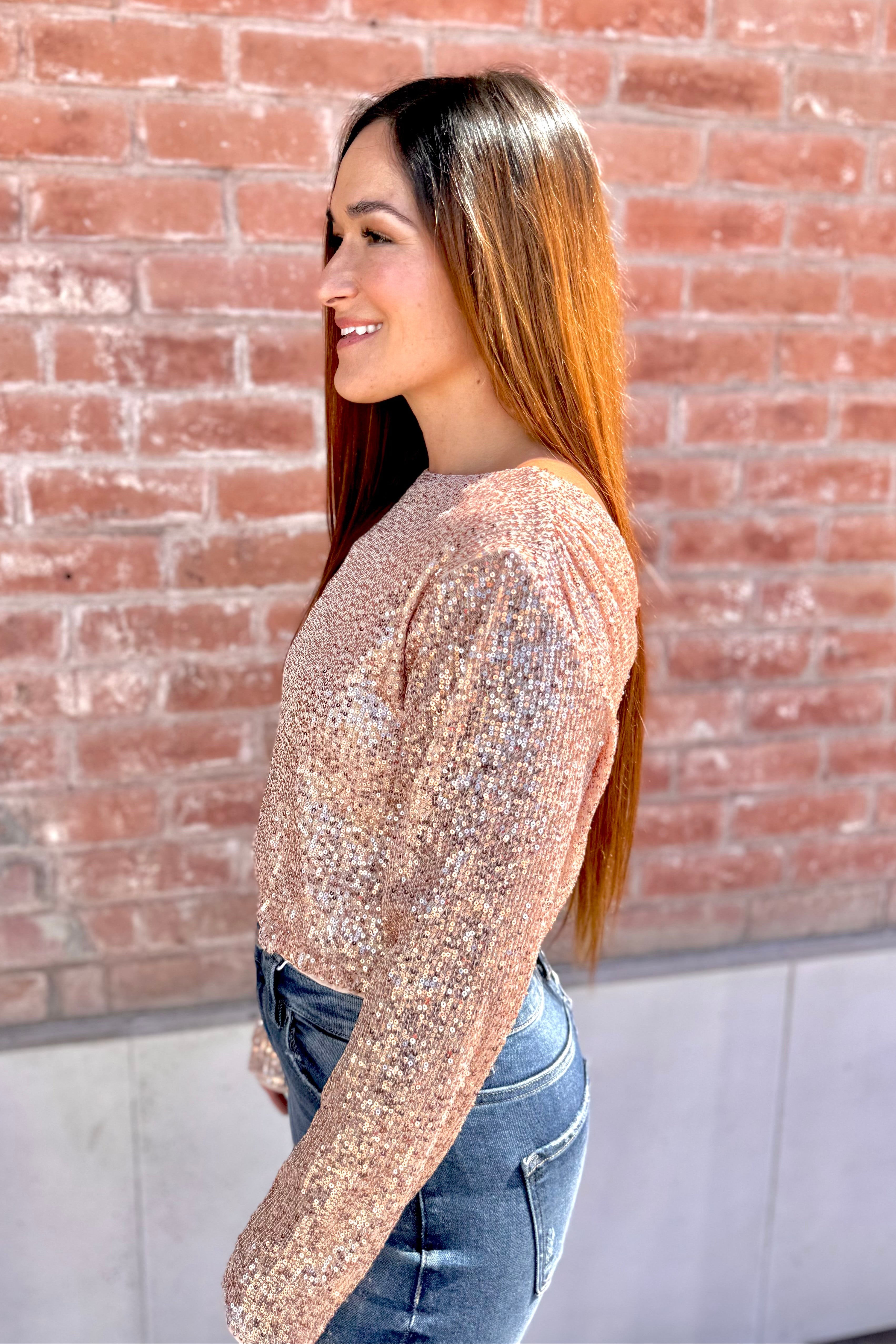 Woman wearing a sparkly long-sleeve top and jeans standing against a brick wall.