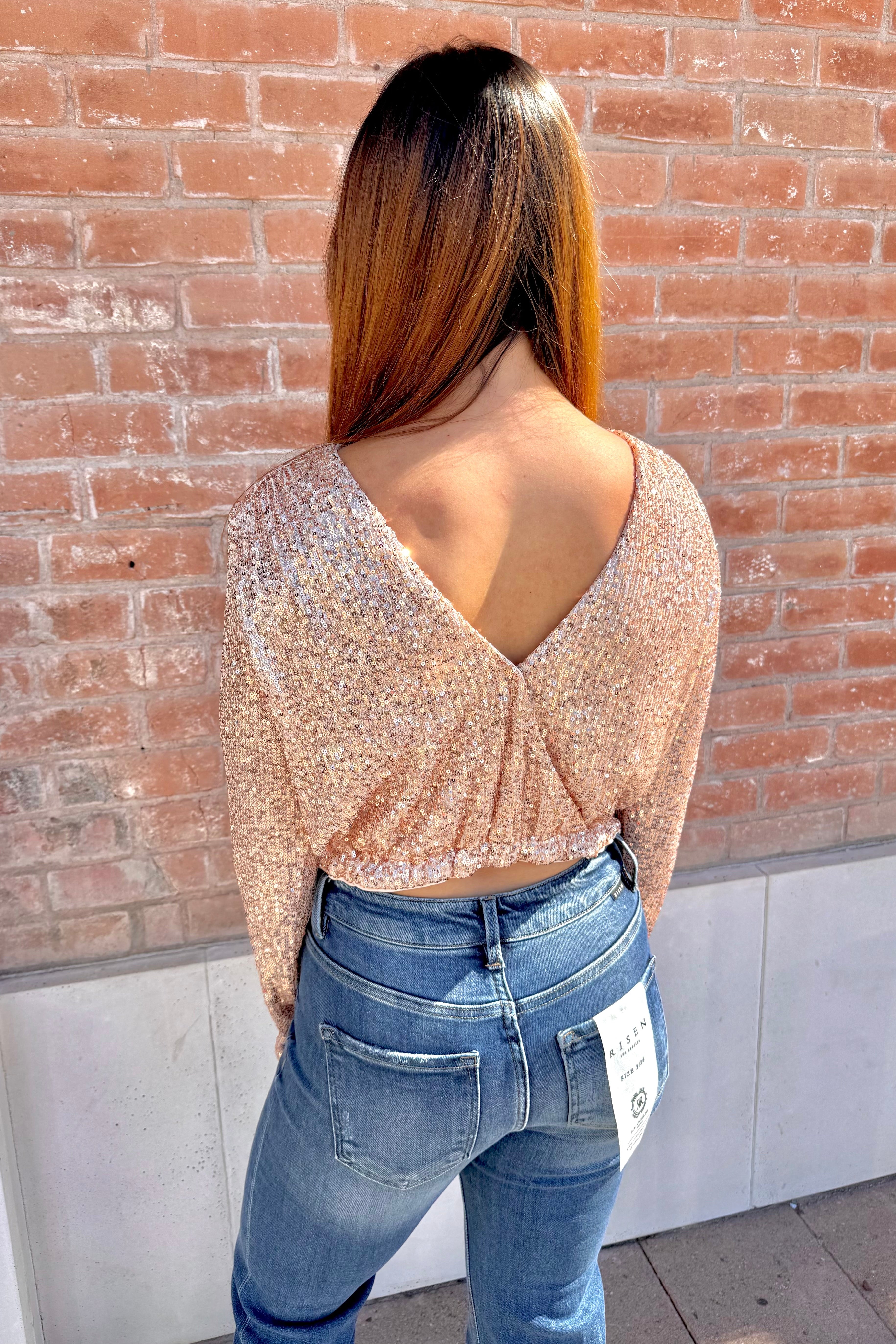 Person wearing a sparkly top and blue jeans against a brick wall.