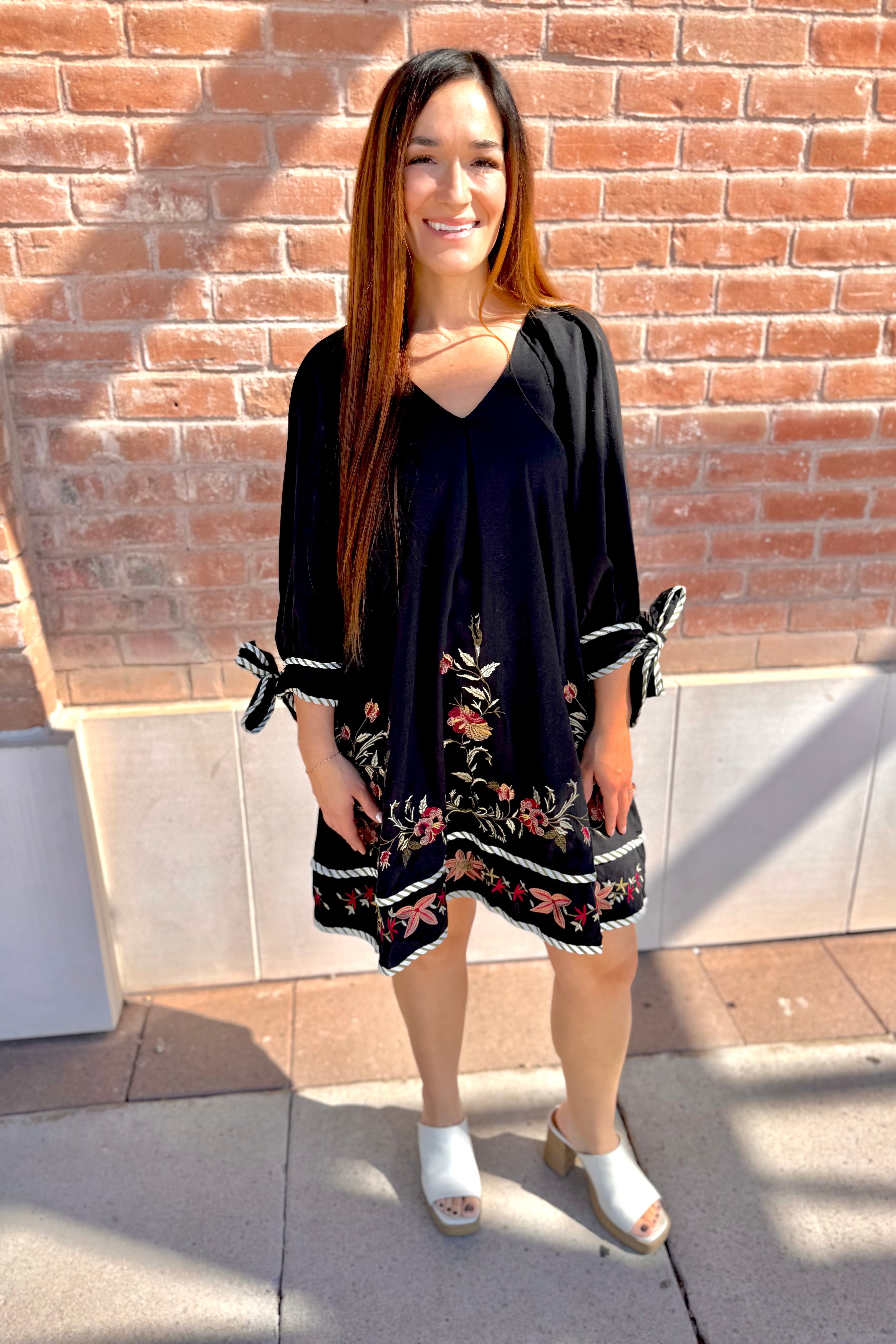 Woman wearing a black dress with floral embroidery standing against a brick wall.