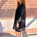 Woman wearing a black dress with butterfly embroidery against a brick wall.