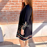 Woman wearing a black dress with butterfly embroidery against a brick wall.