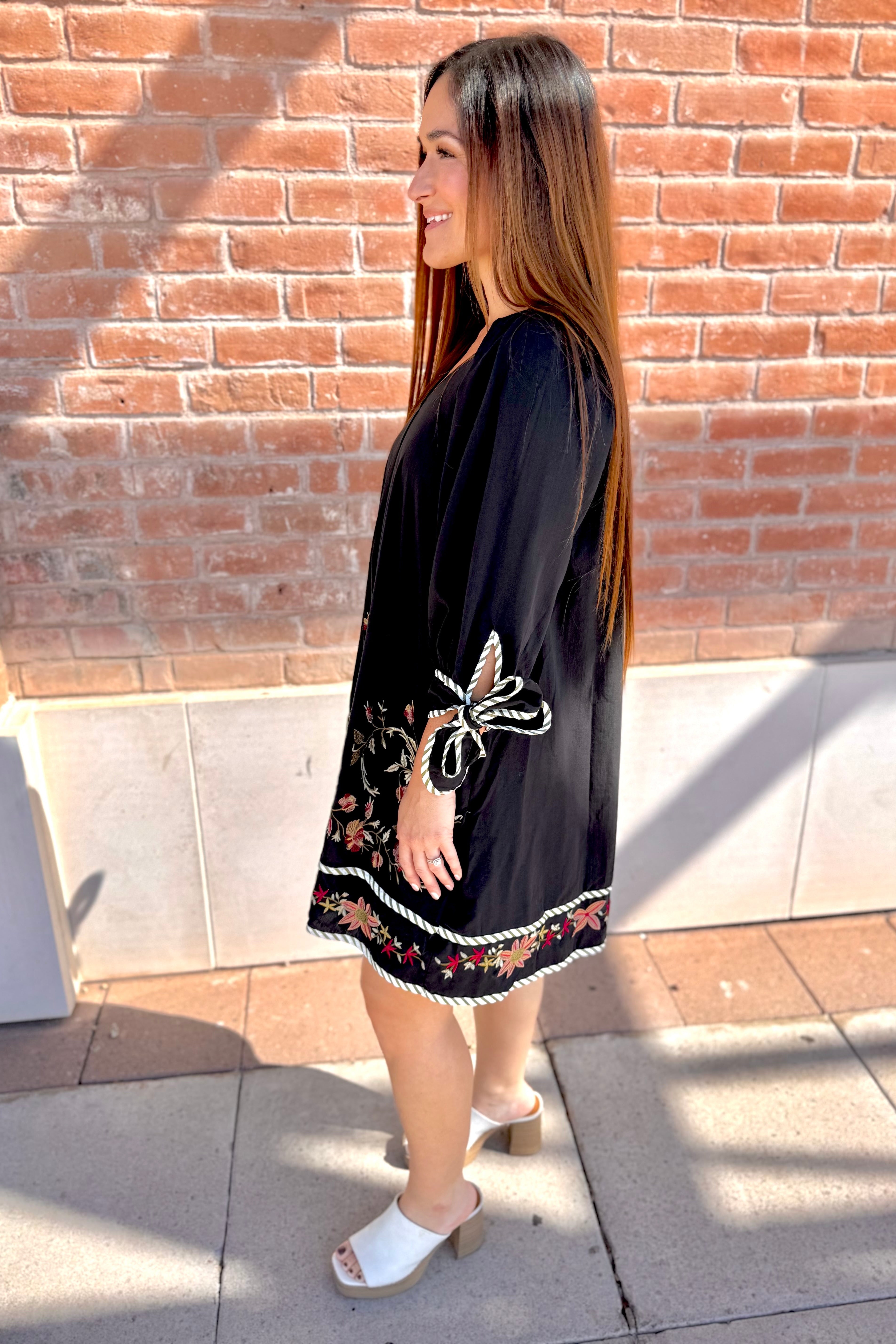 Woman wearing a black dress with butterfly embroidery against a brick wall.
