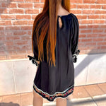 Person wearing a black dress with floral embroidery standing against a brick wall.