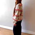 Woman wearing a floral sweater and dark pants standing against a white wall.
