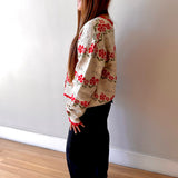 Woman wearing a floral sweater and dark pants standing against a white wall.