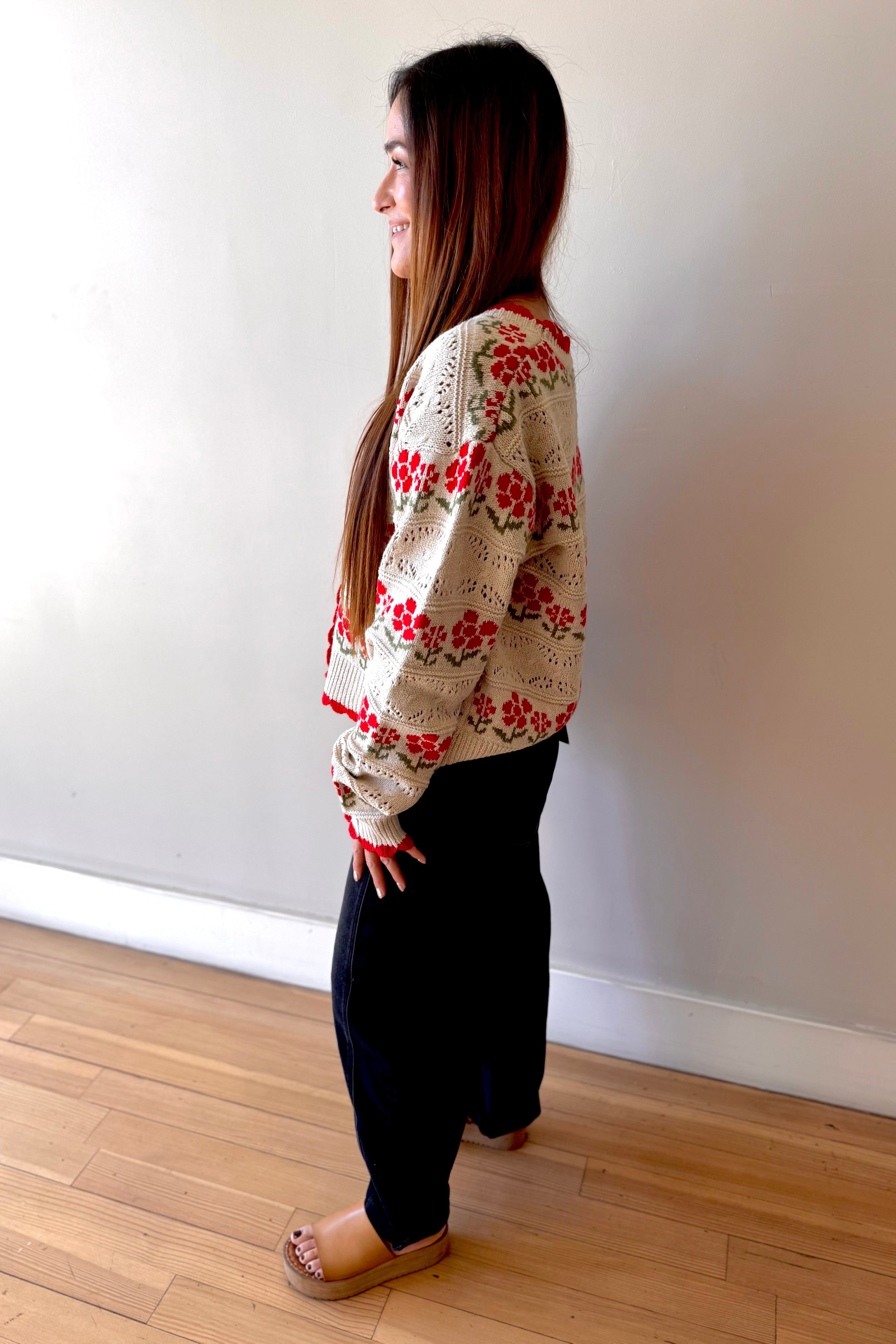 Woman wearing a floral sweater and dark pants standing against a white wall.