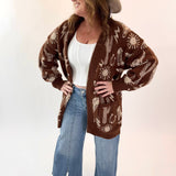 Woman wearing a brown patterned cardigan, white top, blue jeans, and beige hat on a white background