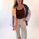 Woman wearing a colorful kimono-style jacket over a brown top and beige pants on a white background
