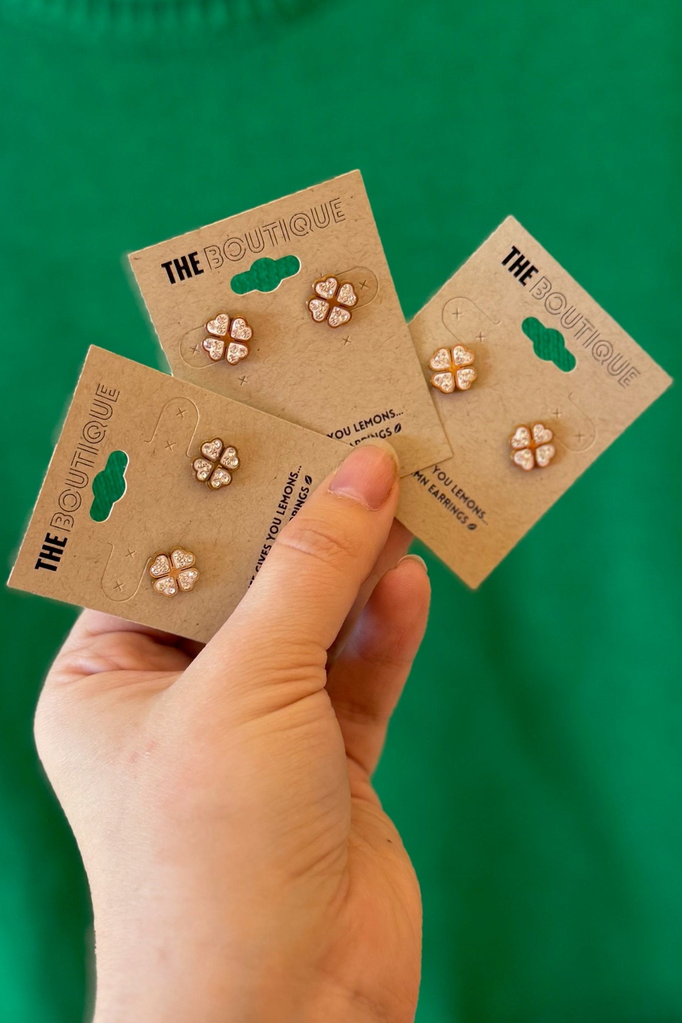 Three pairs of earrings on 'The Boutique' cards held by a hand against a green background