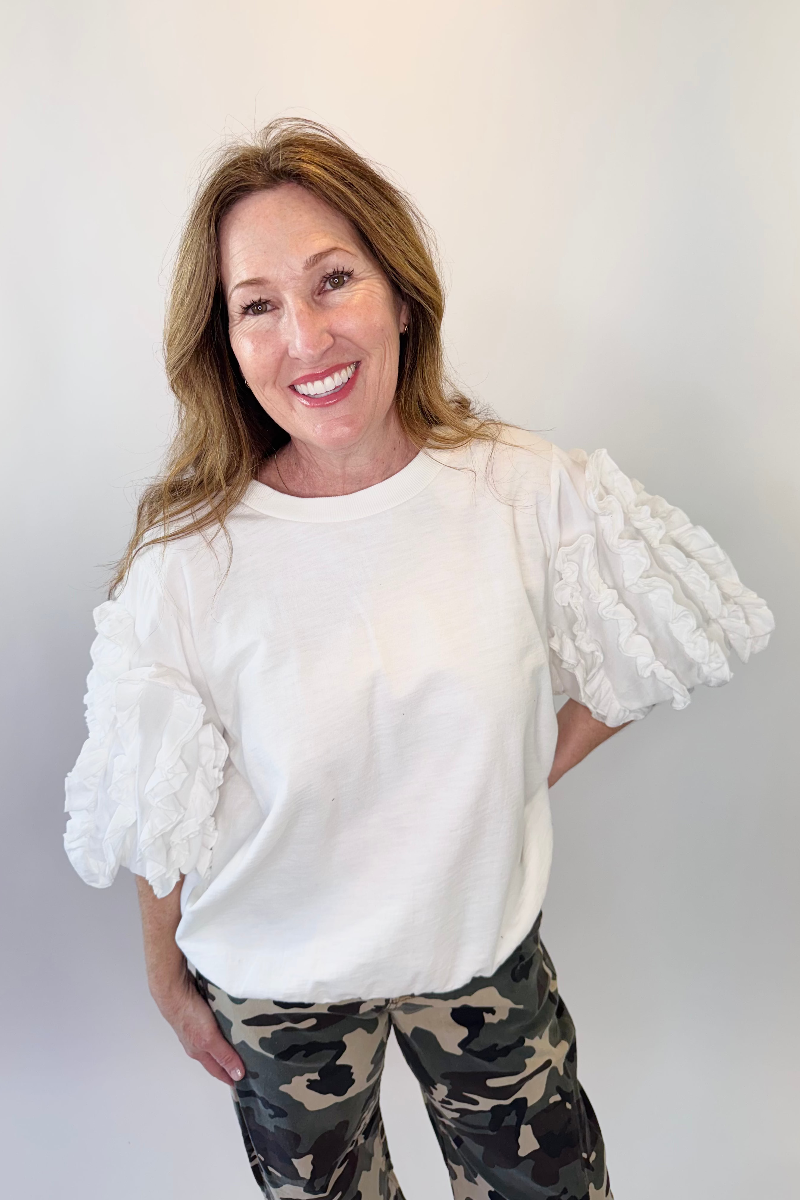 Woman wearing a white blouse with ruffled sleeves and camouflage pants against a plain background