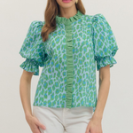 Woman wearing a green patterned blouse with white pants on a plain background