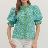 Woman wearing a green patterned blouse with white pants on a plain background