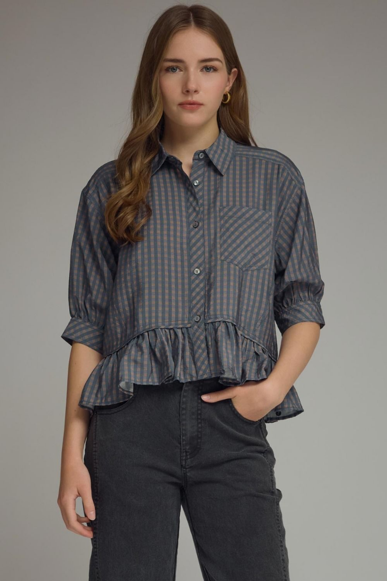Woman wearing a checkered blouse with ruffled bottom and dark pants on a gray background