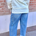 Person wearing a light-colored top and blue jeans standing against a brick wall.