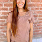 Woman wearing a brown t-shirt and blue jeans standing against a brick wall.