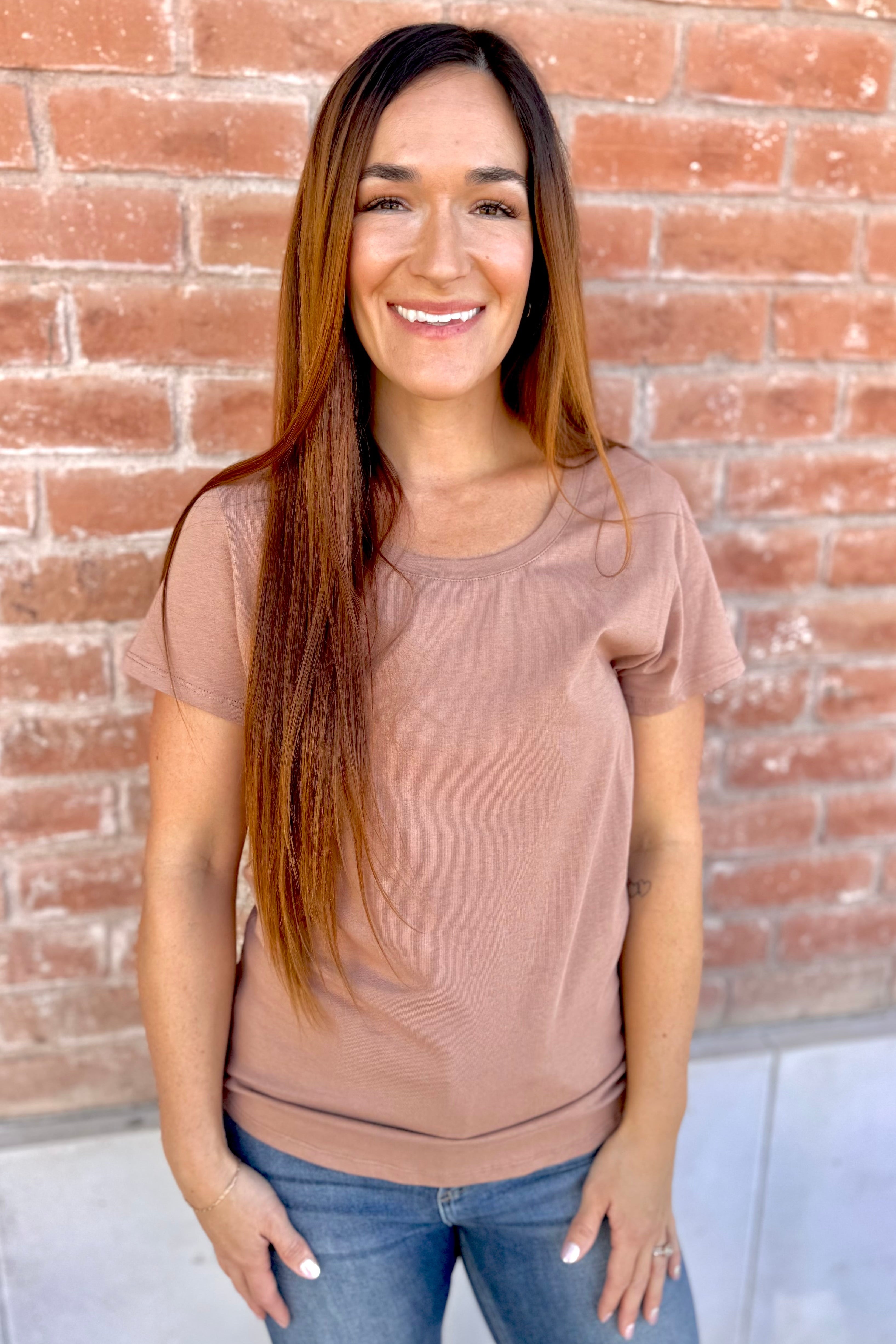 Woman wearing a brown t-shirt and blue jeans standing against a brick wall.