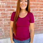Woman wearing a maroon shirt and blue jeans standing in front of a brick wall.