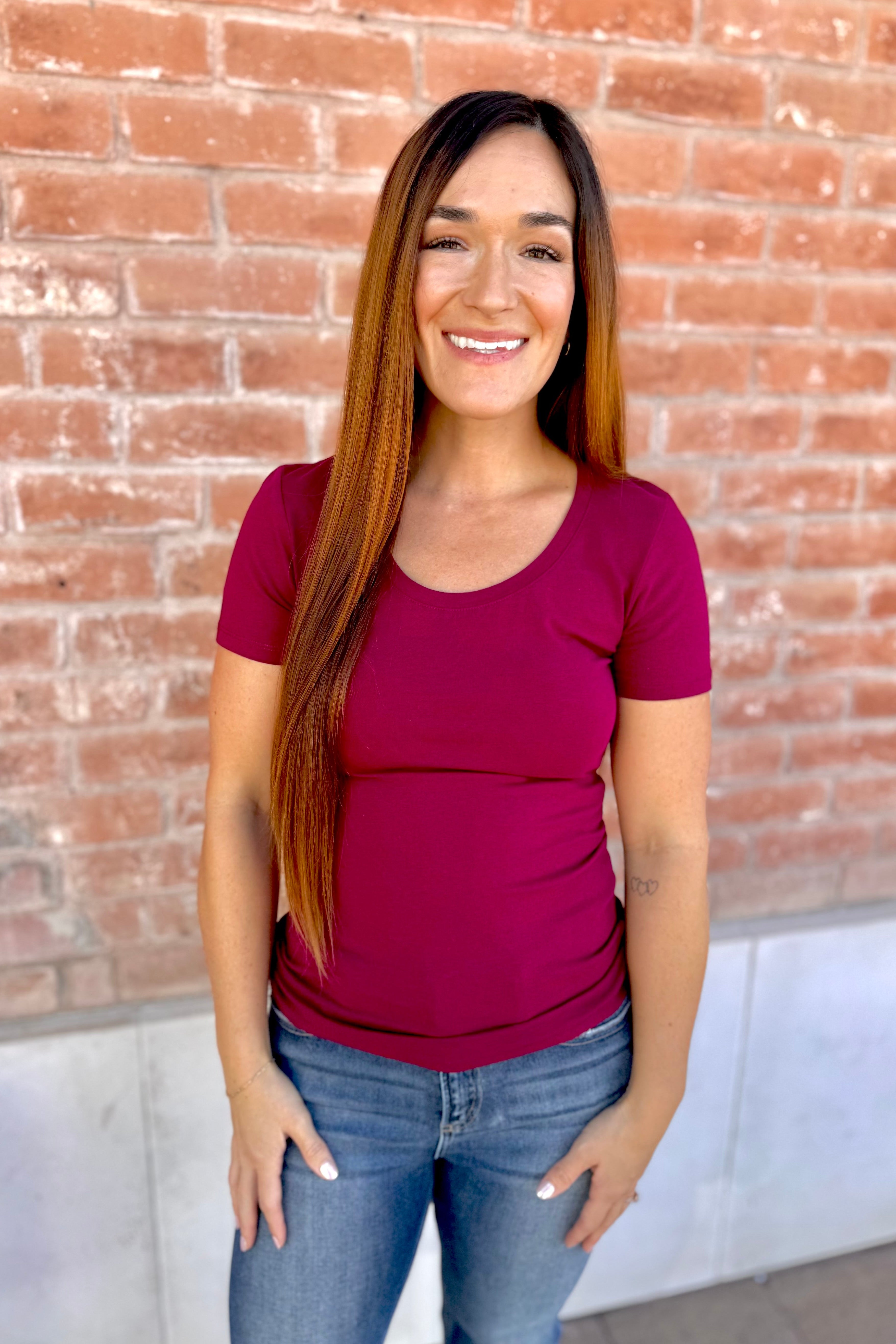 Woman wearing a maroon shirt and blue jeans standing in front of a brick wall.