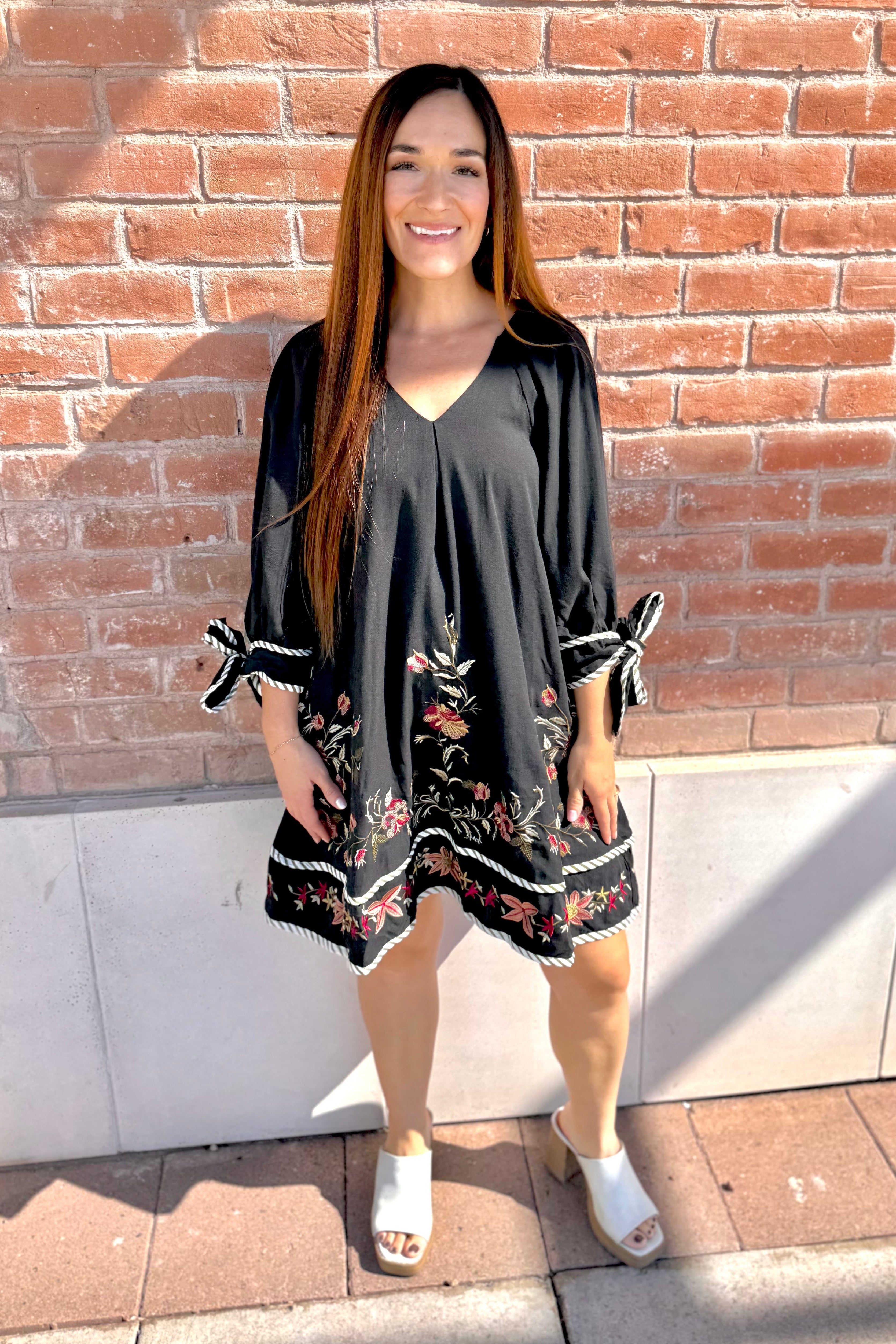 Woman wearing a black dress with floral patterns standing against a brick wall.