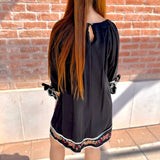 Person wearing a black dress with floral embroidery standing against a brick wall.