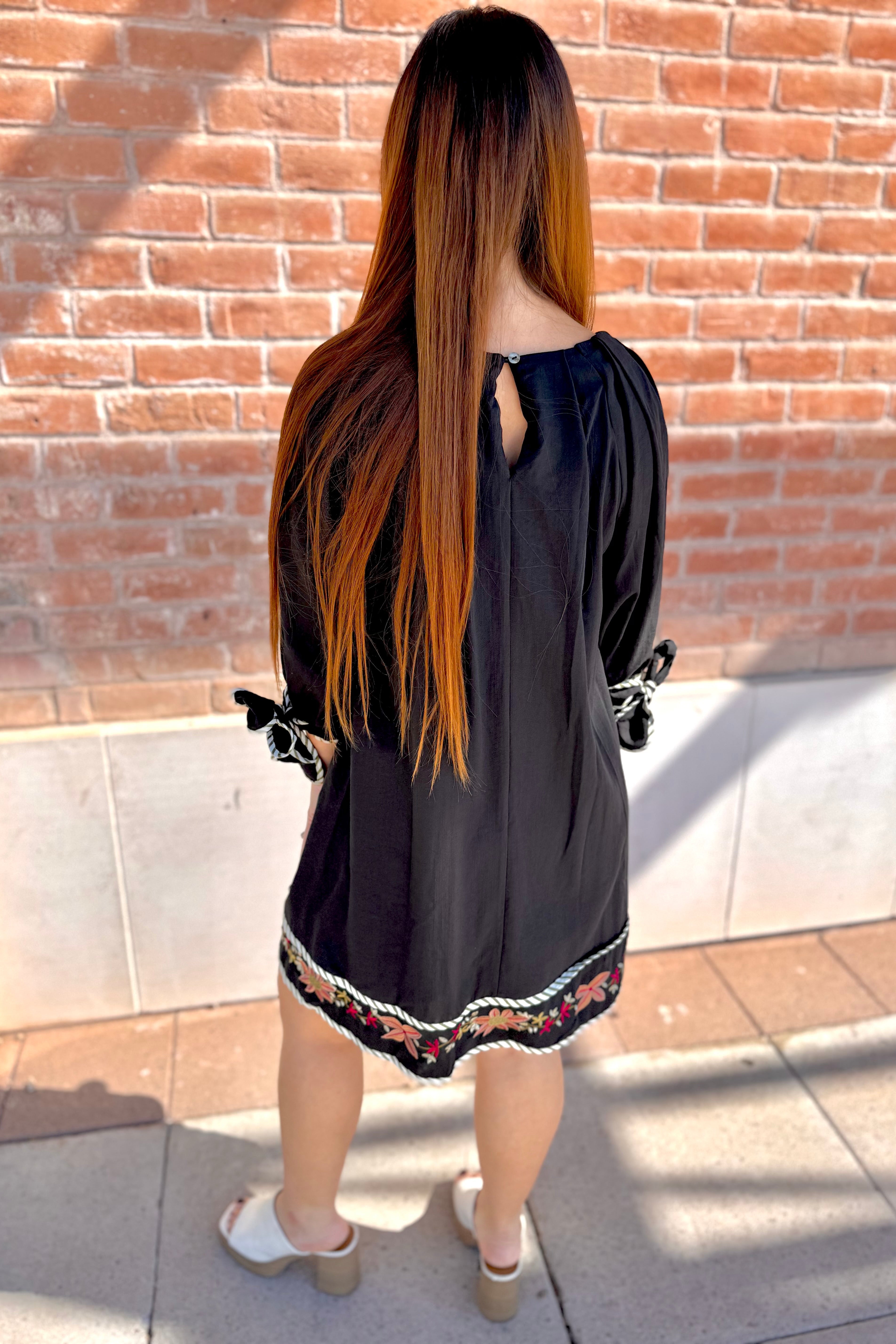 Person wearing a black dress with floral embroidery standing against a brick wall.