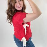 Woman wearing a red sweater with a white bow and blue jeans against a plain background