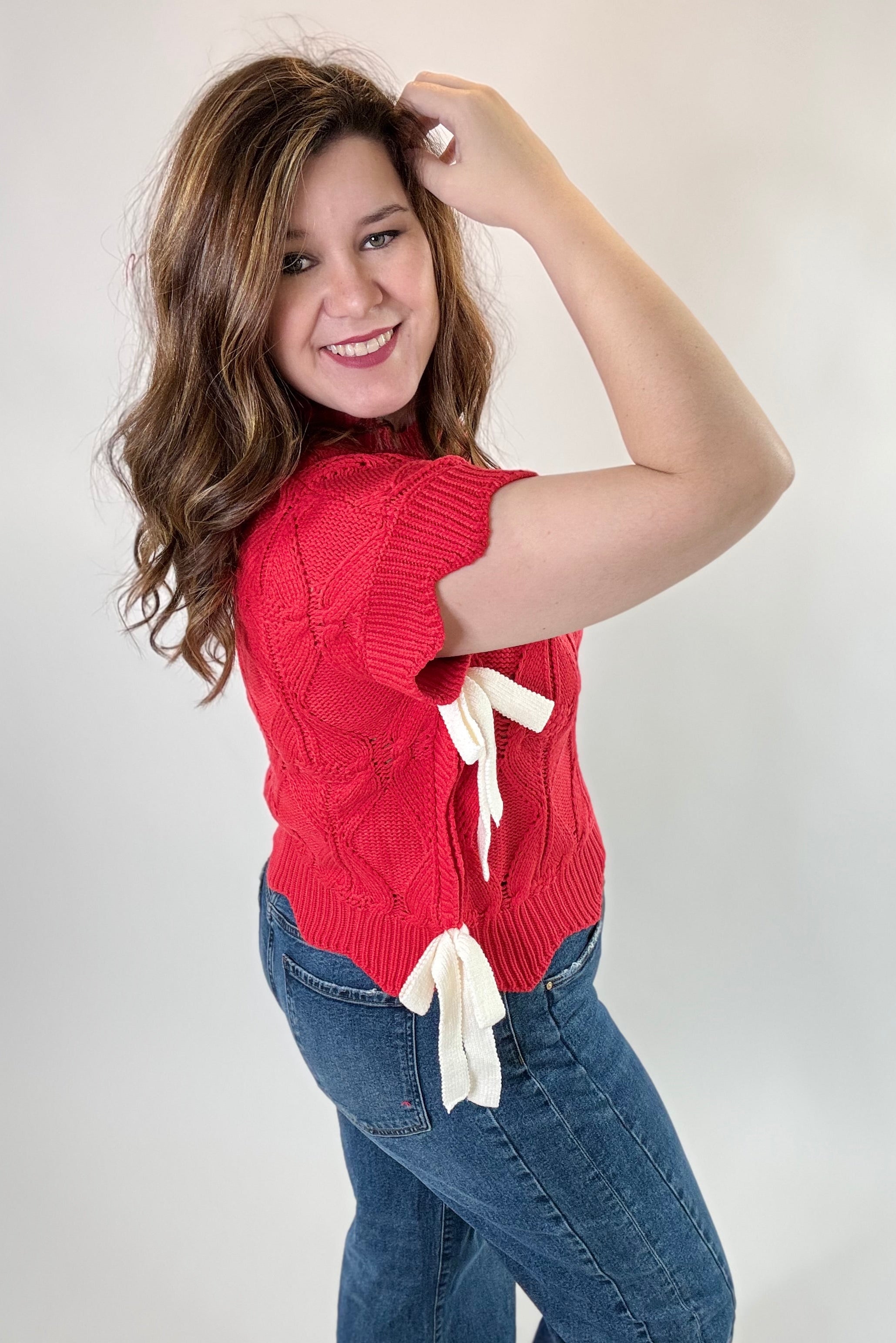 Woman wearing a red sweater with a white bow and blue jeans against a plain background