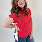 Woman wearing a red sleeveless sweater and blue jeans against a plain background