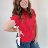 Woman wearing a red sleeveless sweater and blue jeans against a plain background