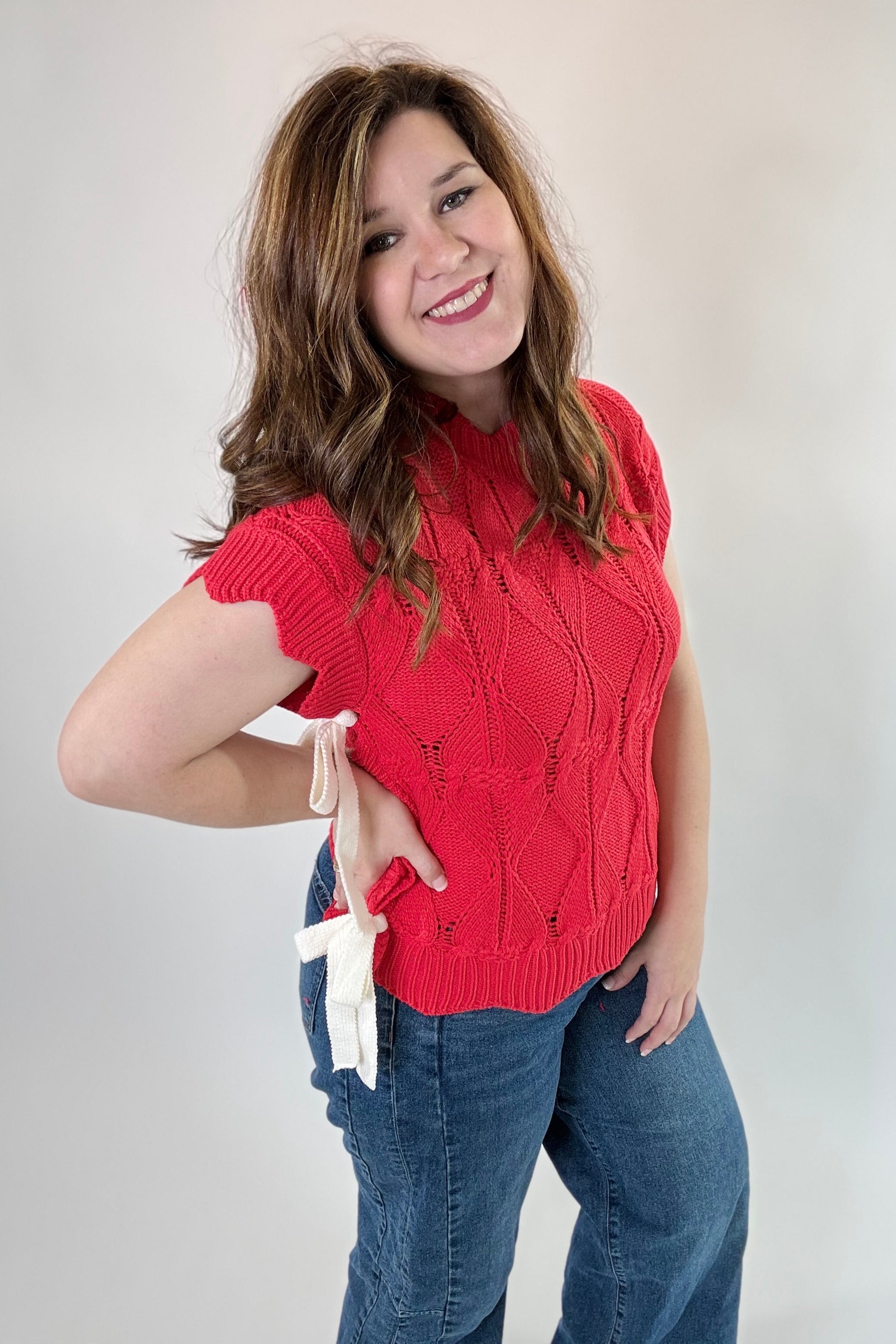 Woman wearing a red sleeveless sweater and blue jeans against a plain background