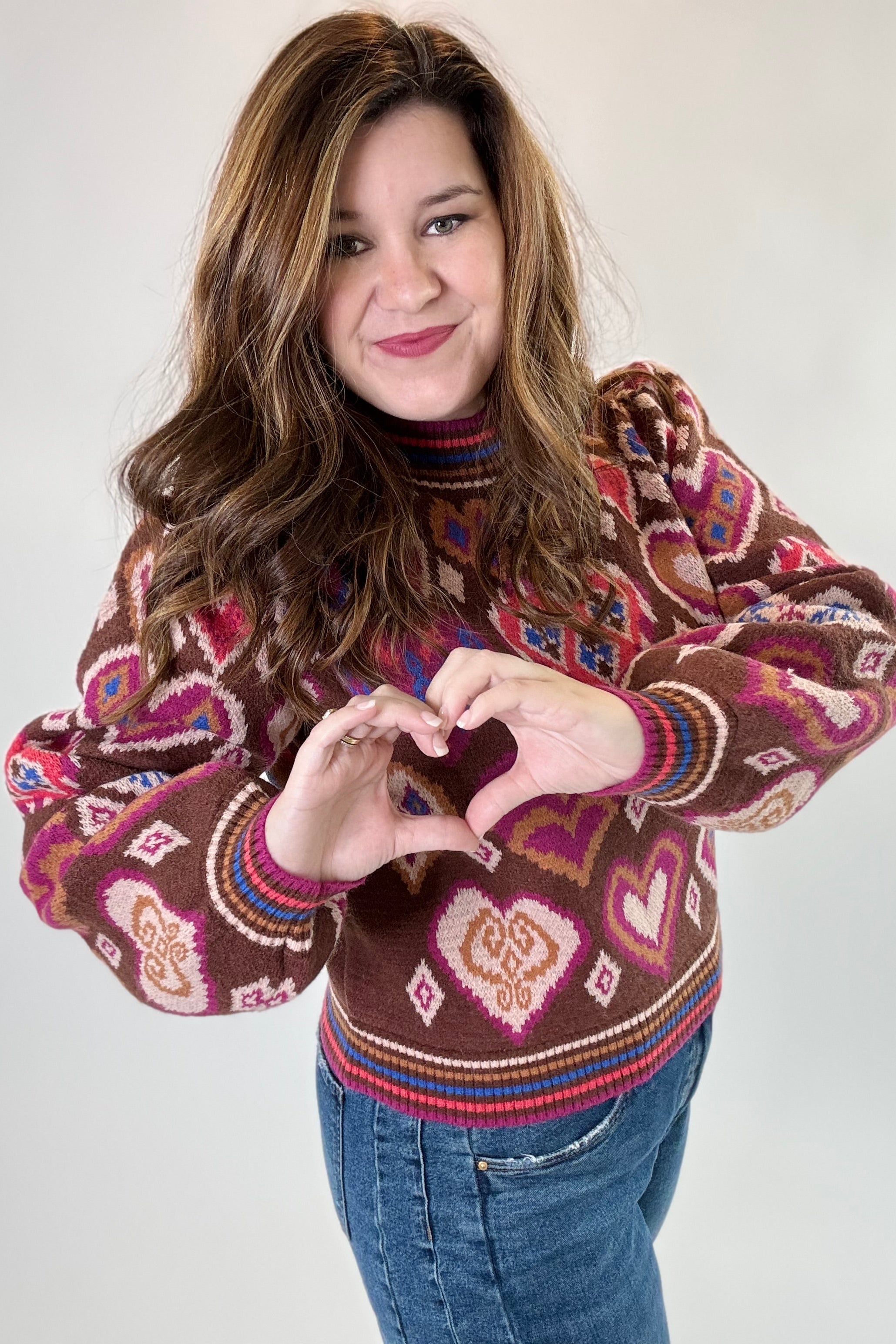 Person wearing a patterned sweater making a heart shape with their hands on a plain background
