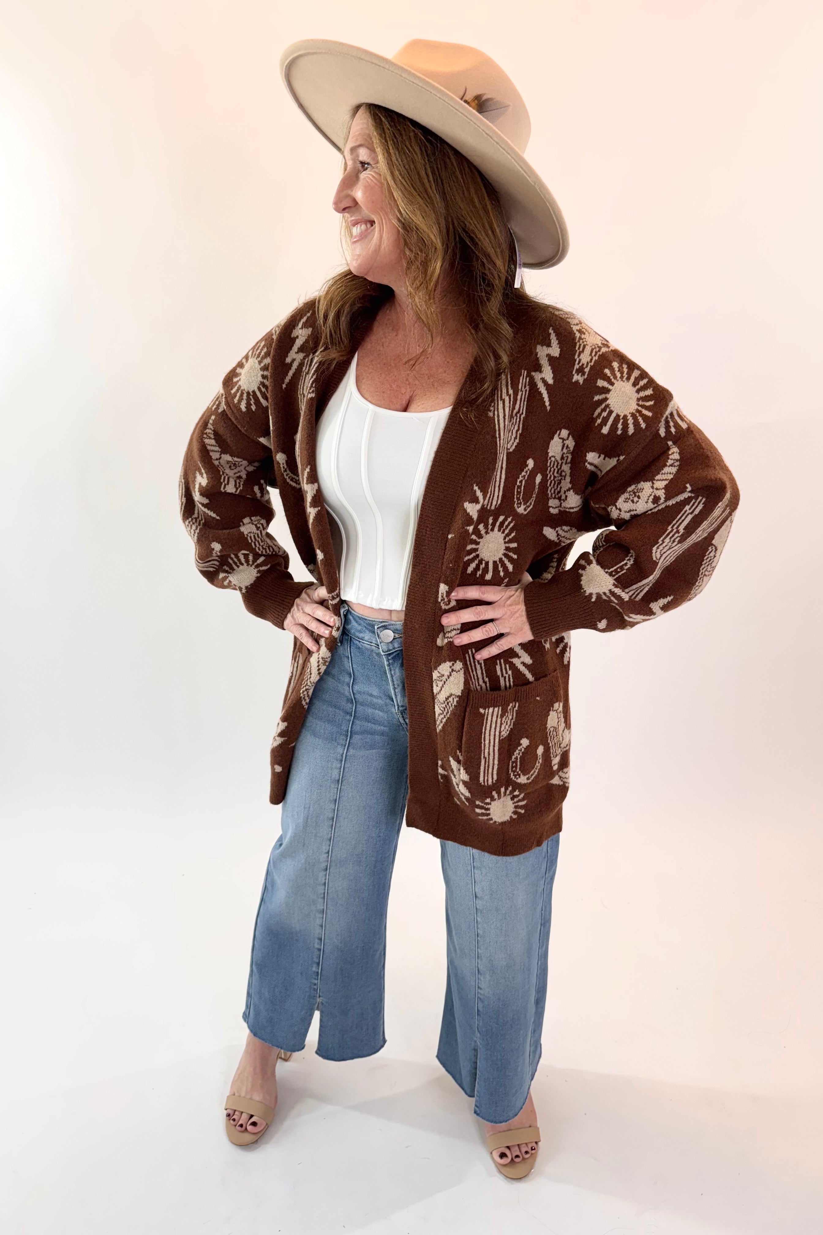 Woman wearing a brown patterned cardigan, white top, blue jeans, and beige hat on a white background