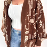 Woman wearing a brown cardigan with white patterns, white top, and blue jeans on a white background