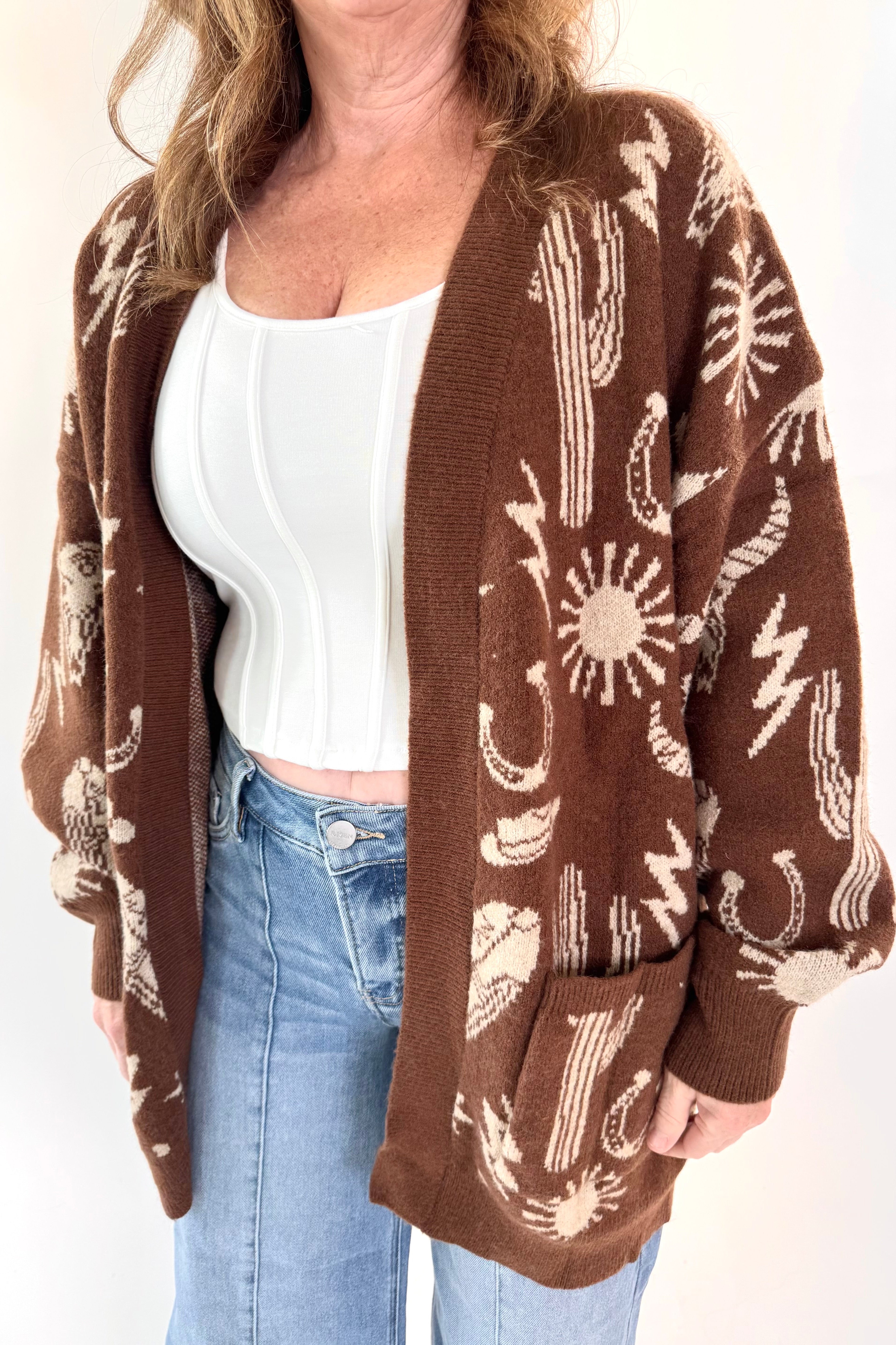 Woman wearing a brown cardigan with white patterns, white top, and blue jeans on a white background