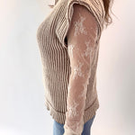 Person wearing a beige ribbed sweater over a sheer lace top with blue jeans on a white background