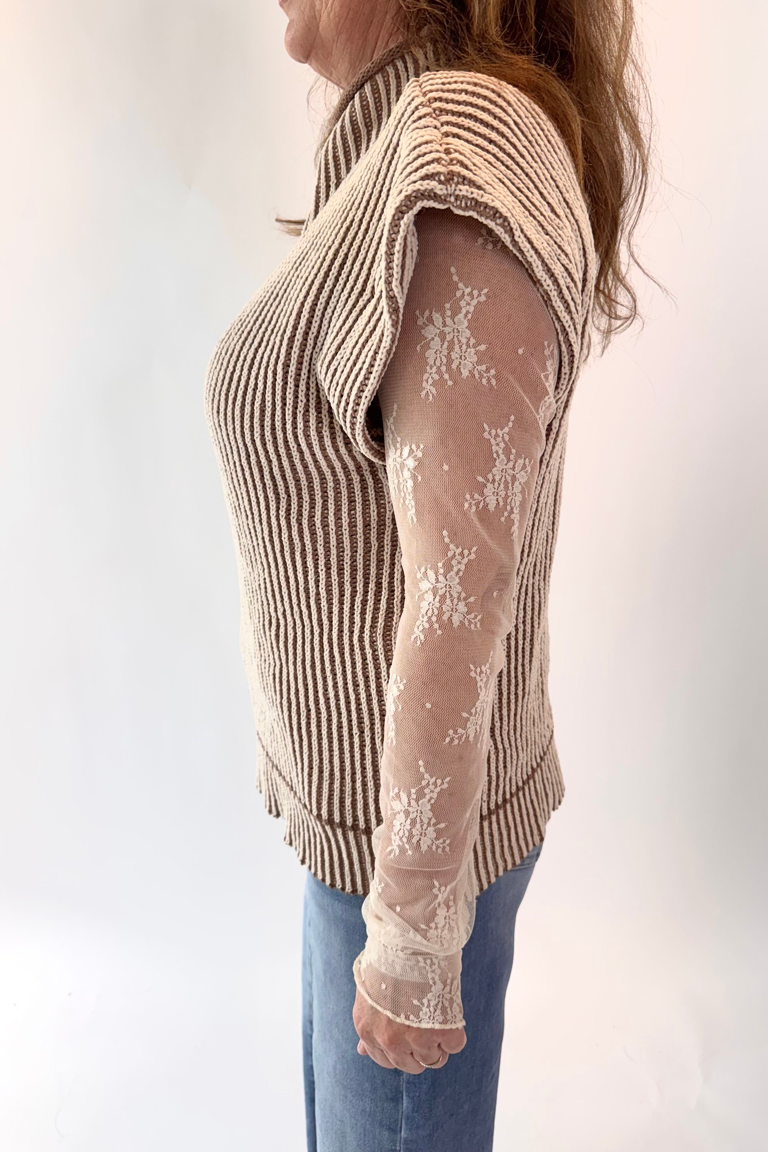 Person wearing a beige ribbed sweater over a sheer lace top with blue jeans on a white background