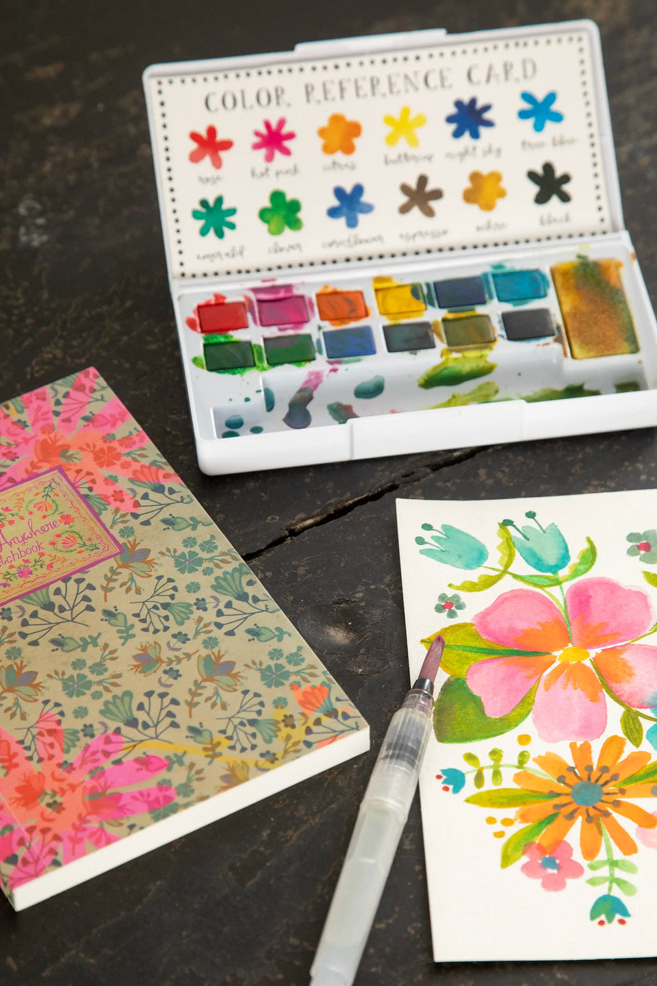 Watercolor paint set with a color preference card, notebook, and watercolor painting on a dark surface.