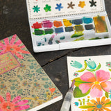 Watercolor paint set with a color preference card, notebook, and watercolor painting on a dark surface.