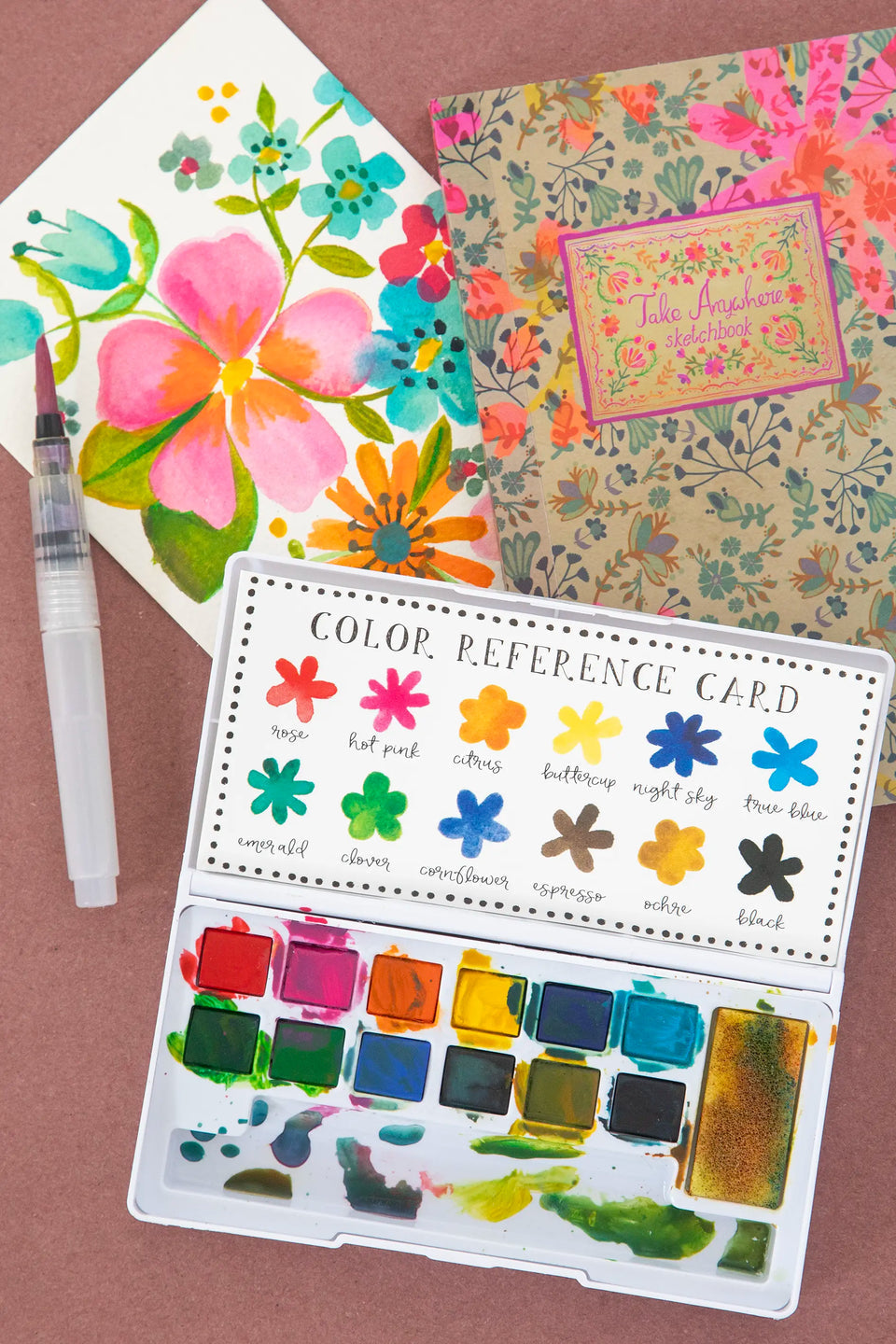 Color reference card with watercolor swatches, floral notebook, and paintbrush on a brown background
