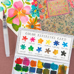 Color reference card with watercolor swatches, floral notebook, and paintbrush on a brown background